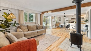 Een ruime woonkamer met een bank, een vloerkleed en een zwarte kachel. Er is een eethoek met stoelen en een tafel.