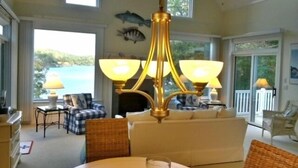Un salón amplio con un sofá blanco grande, una mesa de comedor con un centro de mesa de cristal y una araña. Hay dos ventanas con vistas a un cuerpo de agua y un balcón con mobiliario de exterior.