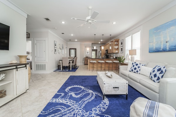 Un salon spacieux avec un grand tapis bleu, un canapé blanc orné de coussins à motifs, une table basse et un ventilateur de plafond.