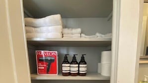 Une salle de bain avec un lavabo blanc, une poubelle blanche et des étagères où sont rangées des serviettes pliées et des articles de toilette.