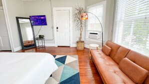 Une chambre à coucher avec un lit, un canapé en cuir marron, une télévision, une plante et une fenêtre avec des stores.