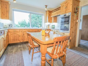 A kitchen with wooden cabinets, a large island with chairs, a Bosch oven, and a window with a view of greenery. - Aelwyd Y Don (Cardigan)
