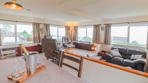 A cozy living room with a sofa, a dining table with glasses and a bottle, and a view of the outdoors through large windows. - Aelwyd Y Don (Cardigan)