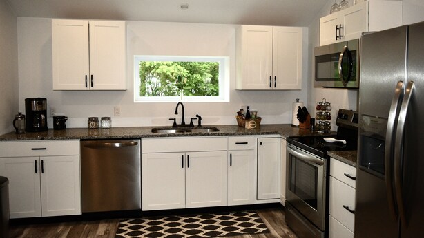 A modern kitchen with white cabinets, stainless steel appliances, a window with a view of greenery, and a patterned rug. - Renovated Modern Gem w/AC – Walk to Cooperstown All-Star Village, Sleeps 6 (Oneonta)