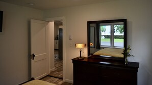 A hallway with a closed door, a lamp on a dresser, and a mirror reflecting a window view. - Renovated Modern Gem w/AC – Walk to Cooperstown All-Star Village, Sleeps 6 (Oneonta)