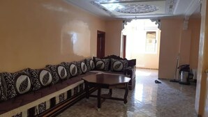 A room with a long, patterned sofa and a wooden table, a tiled floor, and a decorative ceiling. - Family apartment in Tagadirte! (Agadir)