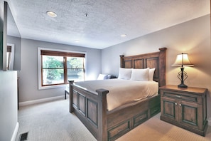 A bedroom with a large bed, a nightstand with a lamp, a window with blinds, and a ceiling with recessed lighting. - Townhouse right off Park City's Rail Trail| Managed by Park City Lodging (Park City)
