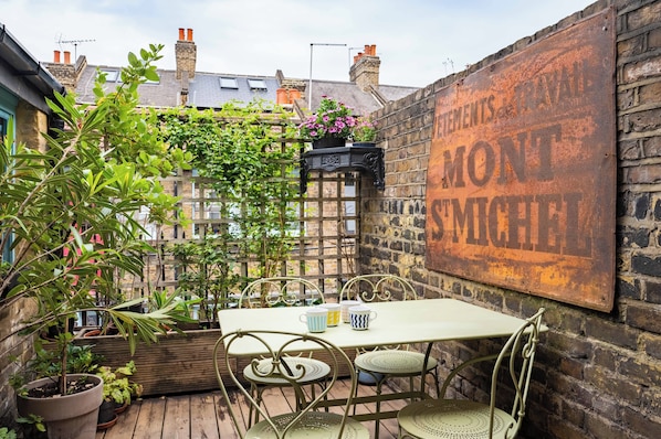 Relax in a charming outdoor seating area with a metal table and four chairs, surrounded by lush greenery and a wooden deck.