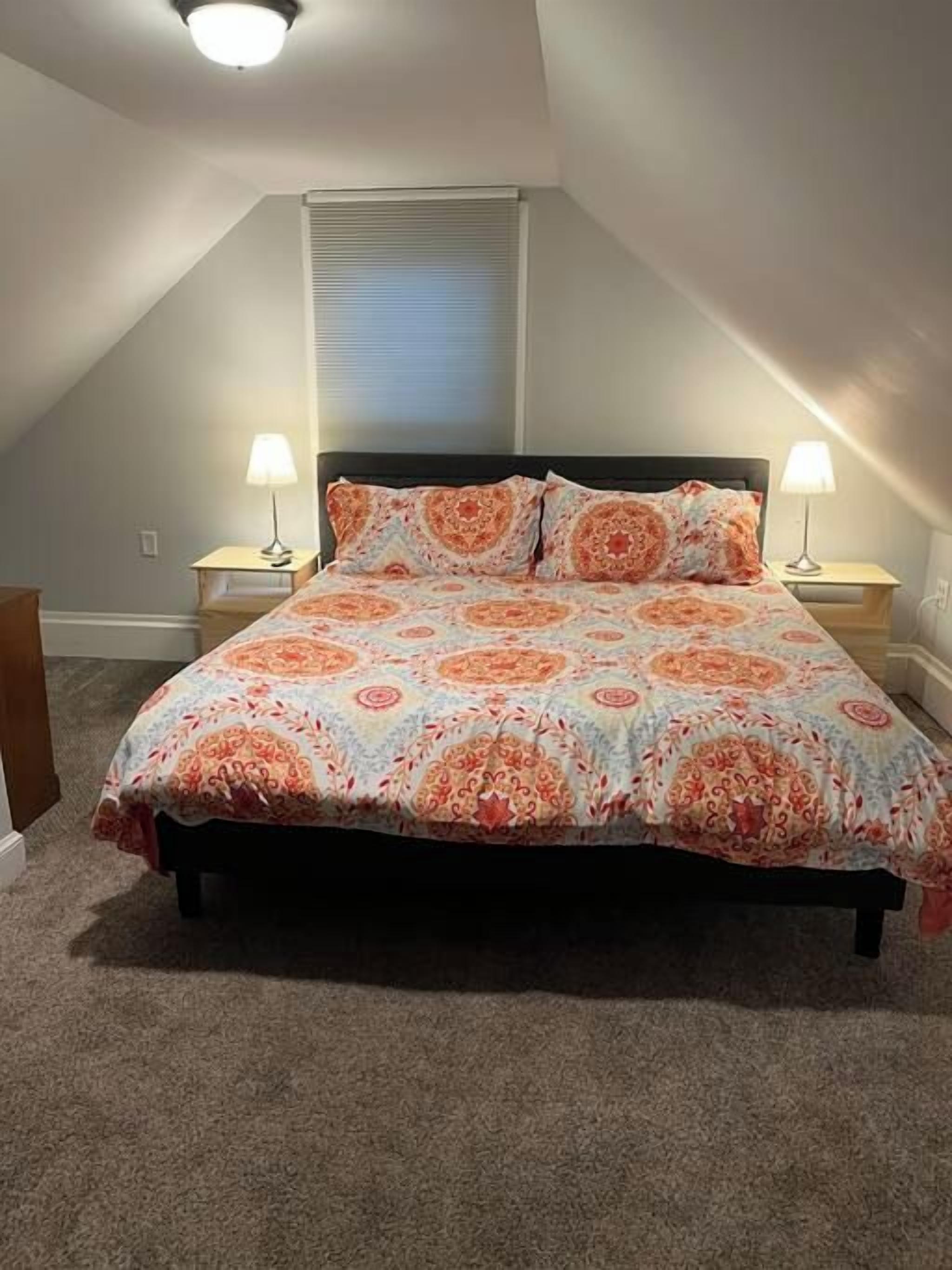 A bedroom with a bed, two bedside lamps, a window with blinds, and a ceiling light.