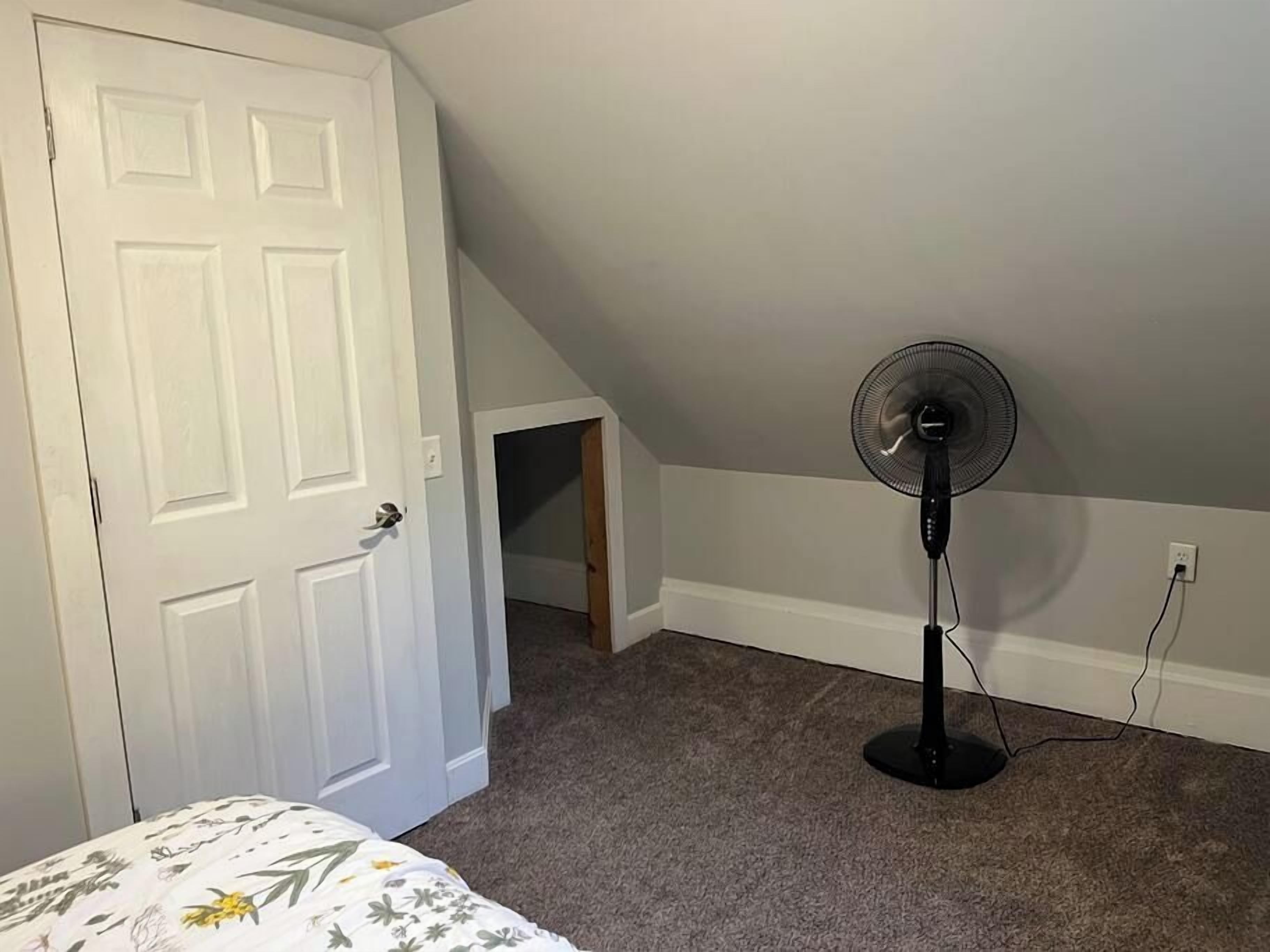 Une pièce avec une porte blanche, un ventilateur sur pied et un sol recouvert de moquette.