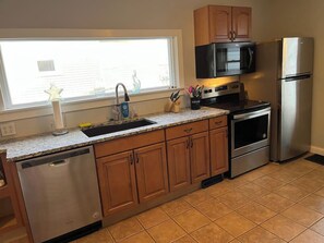 Una cocina con armarios de madera, electrodomésticos de acero inoxidable, un microondas, un refrigerador y un ventanal con persianas.