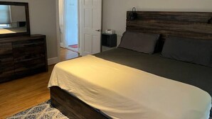 A bedroom with a wooden headboard, a large bed with white bedding, a dresser with a mirror, and a door leading to another room. - Main, Three Bedroom House in Historic Loveland (Loveland)