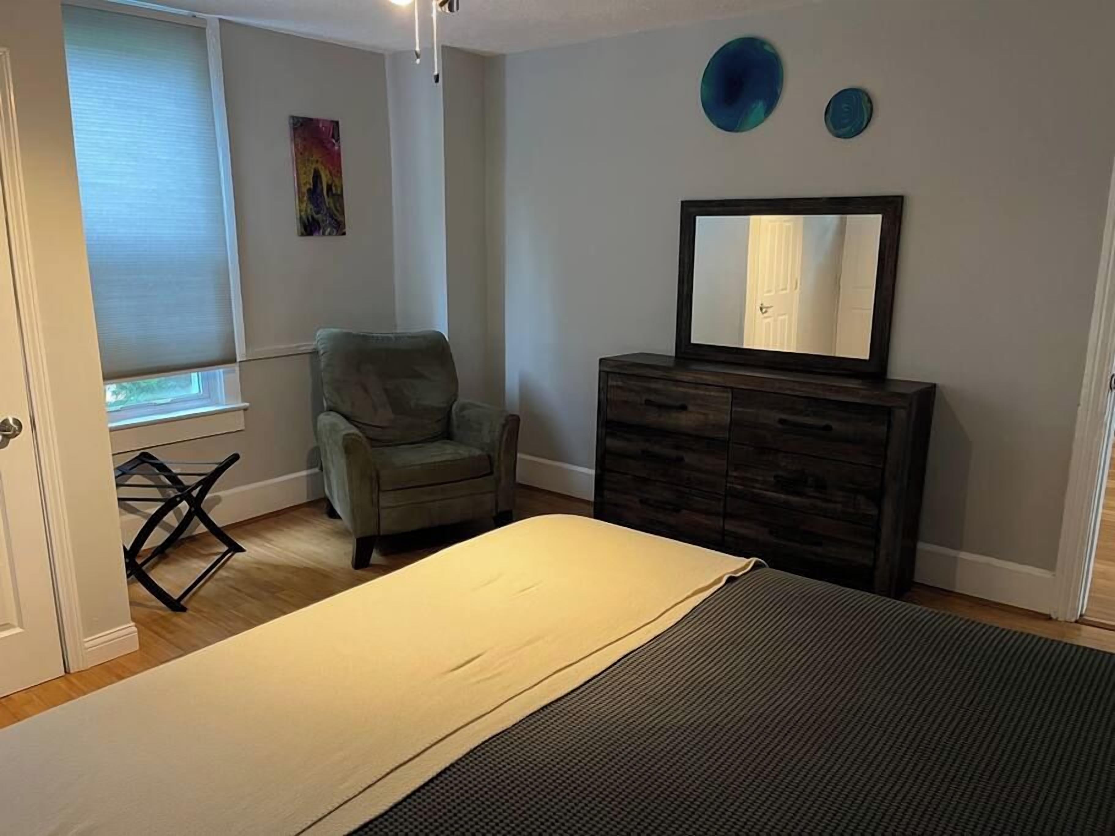 Quarto com cama, cômoda, cadeira, janela com persianas e parede com duas pratos decorativas.