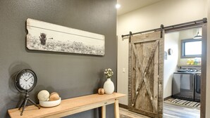 Une console en bois ornée d’une horloge et d’objets décoratifs, une porte de grange rustique et, au fond, une cuisine.