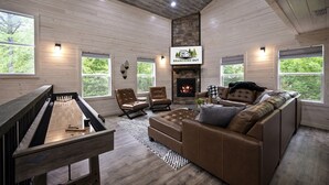 A spacious living room with a fireplace, wooden walls, and large windows offering a view of the outdoors. - Forest Retreat Hot Tub Slide Arcade Family Fun (Broken Bow)