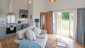 A modern kitchen with a gas stove, microwave, and cabinets. There is a sofa with cushions and a coffee table with a book. The room has a large sliding glass door leading to a patio. - Peep o' Day (York)