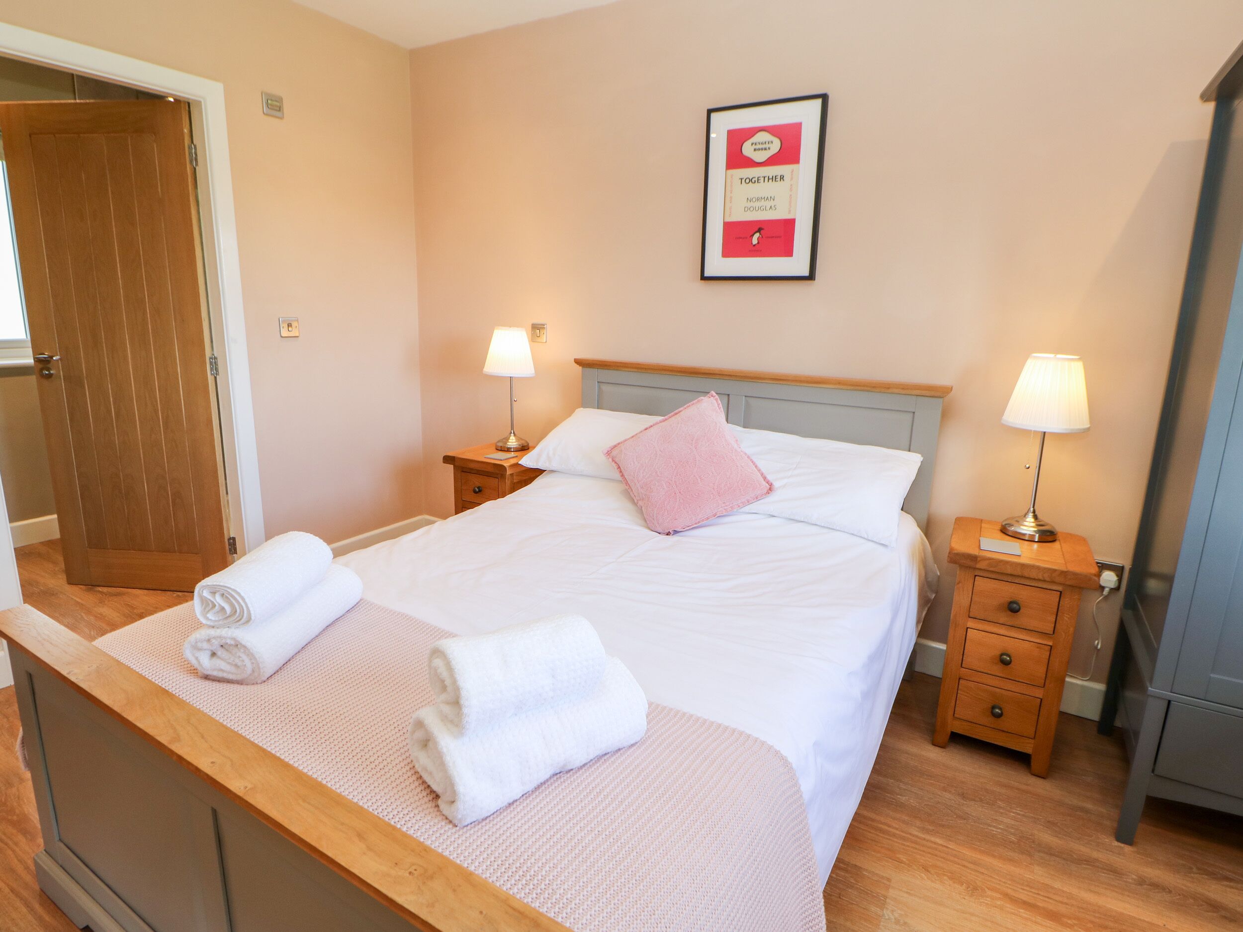 A neatly made bed with white linens and pink pillows, two bedside tables with lamps, a framed book on the wall, and a wooden door.
