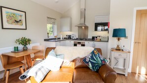 A modern living room with a brown leather sofa, a dining table with chairs, a kitchen area with a stove, and a small side table with a lamp. - Rising Sun (York)