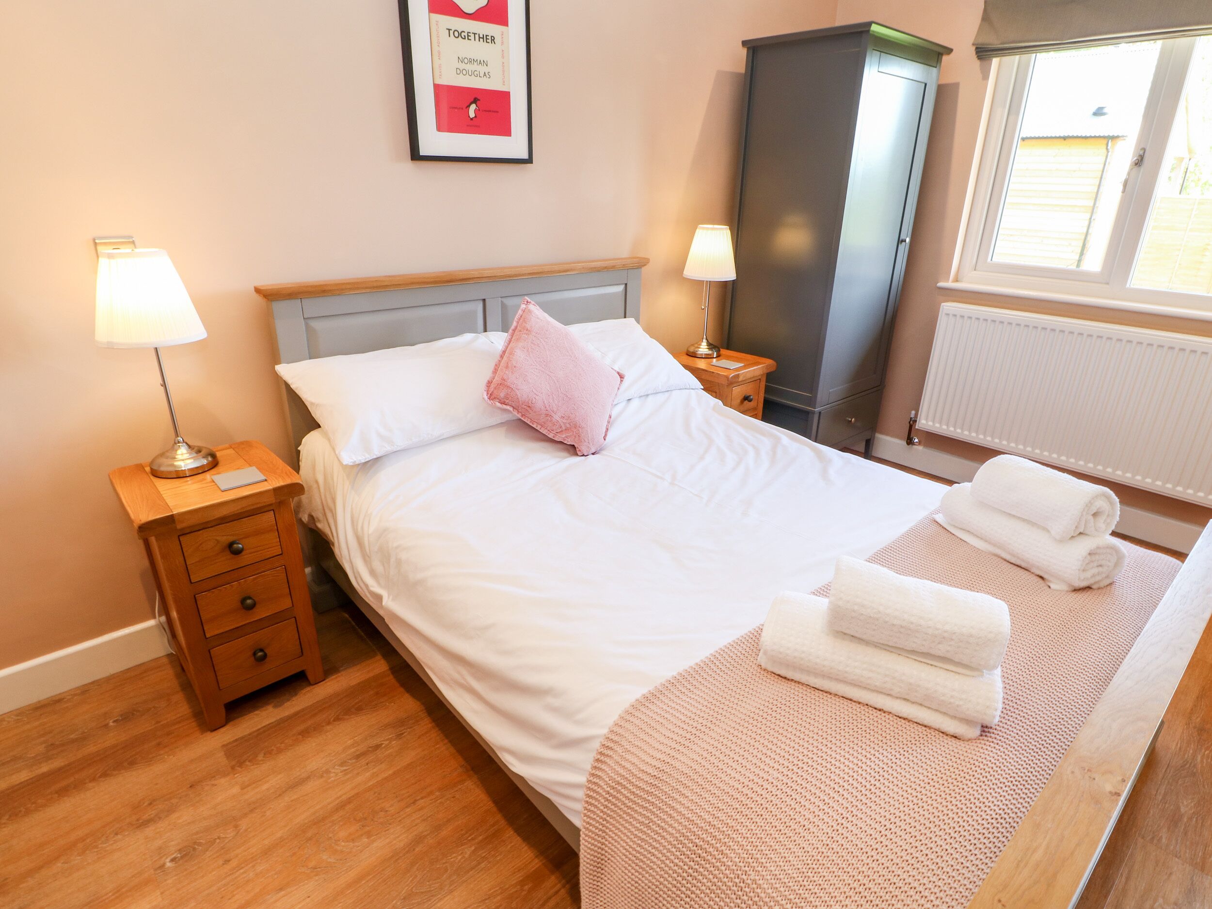 A neatly made bed with white linens and a pink pillow, a wooden nightstand with a lamp, a framed book on the wall, a wardrobe, and a window with daylight.