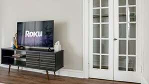 Una sala de estar moderna con un televisor de pantalla plana sobre un mueble negro, una puerta blanca con paneles de vidrio y un piso de madera.