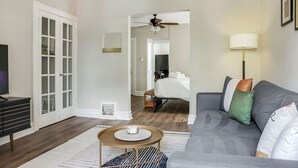Una sala de estar moderna con un sofá gris, una mesa de centro y un televisor. Se observa una puerta con paneles de vidrio y un ventilador de techo.