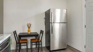 Cocina compacta con refrigerador de acero inoxidable, una pequeña mesa de comedor con dos sillas, y un florero con flores secas sobre la mesa.