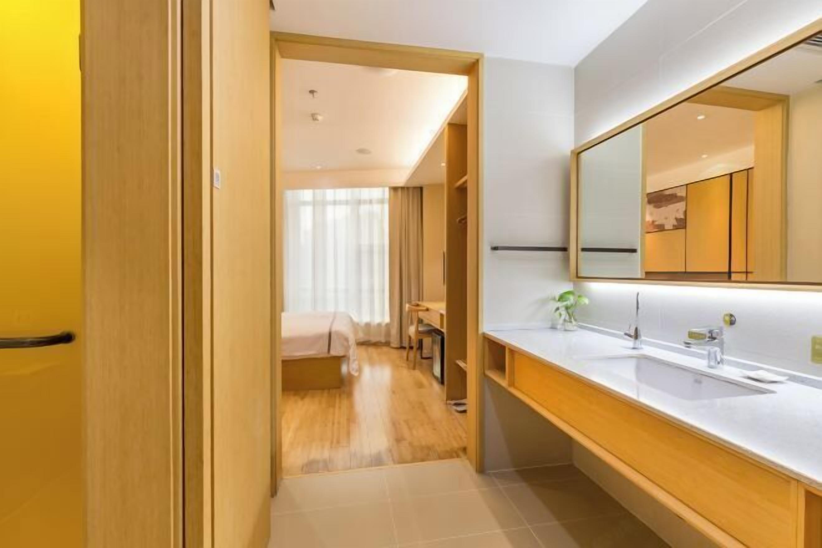 A modern hotel room with a wooden door, a large mirror, a white sink, and a bed with a wooden headboard.