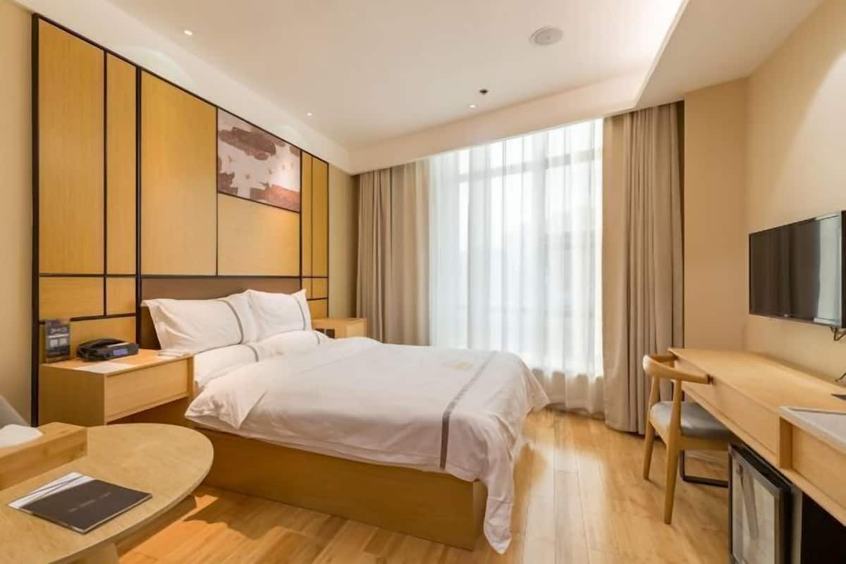 Habitación de hotel con cama, mesitas de noche, escritorio, silla, televisor y un ventanal grande con cortinas.