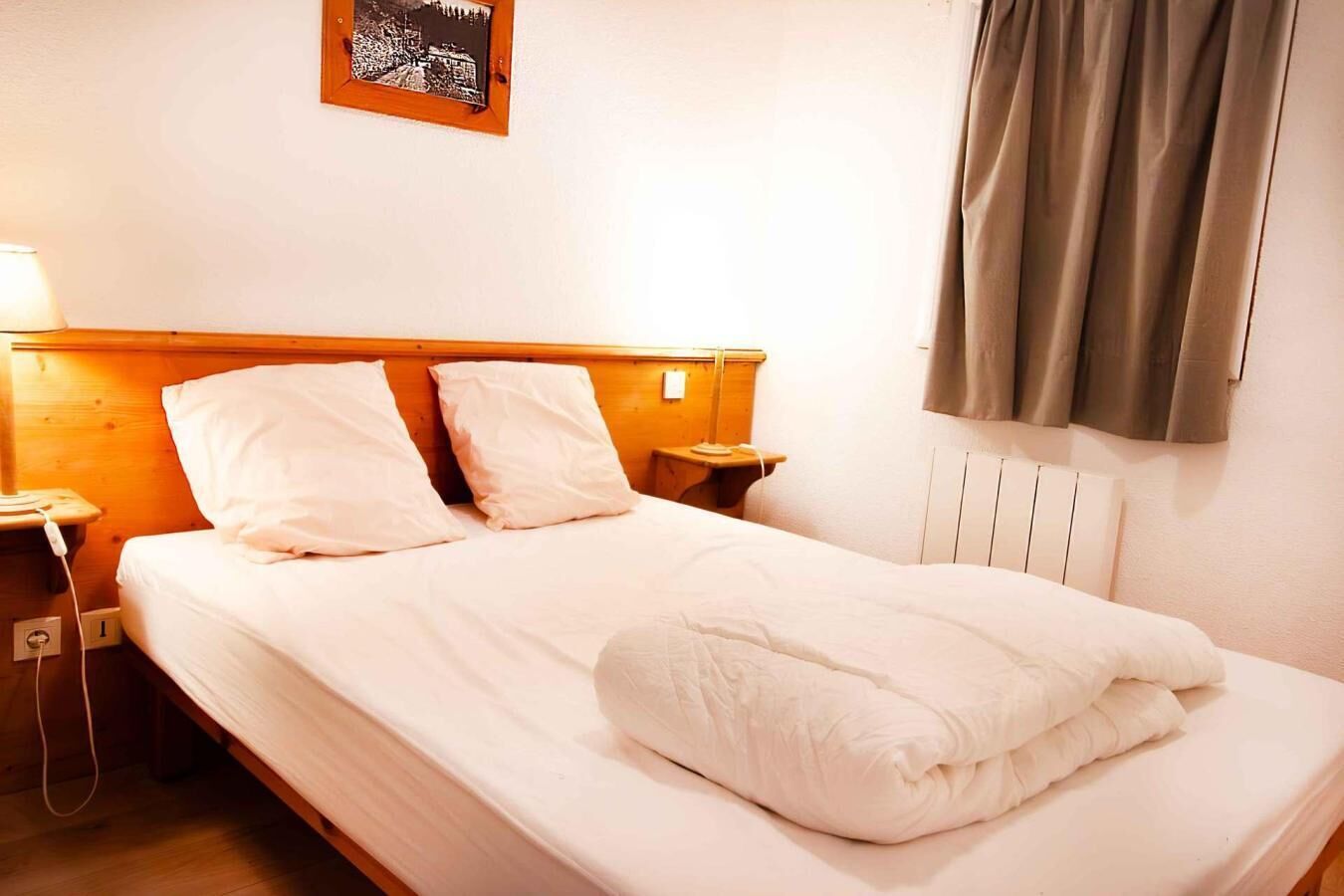 Een net opgemaakt bed met witte lakens en kussens, een houten hoofdbord, een nachtkastje met een lamp en een lijst met een afbeelding aan de muur.
