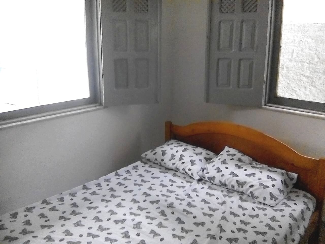 A single bed with a patterned bedspread and two pillows, a wooden headboard, and a window with a view of a building outside.