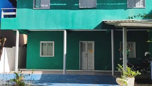 Un bâtiment vert avec deux portes et des fenêtres, un espace abrité où se trouve une moto, et une plante au premier plan.