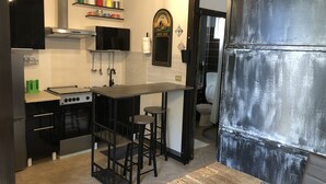 Een kleine keuken met een fornuis, gootsteen en barstoelen. Er zijn planken met bakken en een lijst met een afbeelding aan de muur.