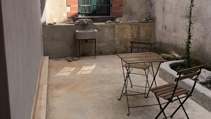Een klein patio buiten met een betonnen vloer, een metalen tafel en twee opklapbare stoelen, een klein spoelgedeelte en een raam met een groen metalen hek.
