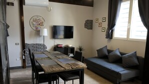 Een woonkamer met een bank, een televisie, een tafel met stoelen, een open haard en een raam met gordijnen.