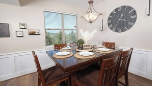 Comedor con mesa de madera dispuesta para cuatro personas, un reloj de pared grande y una ventana con persianas.