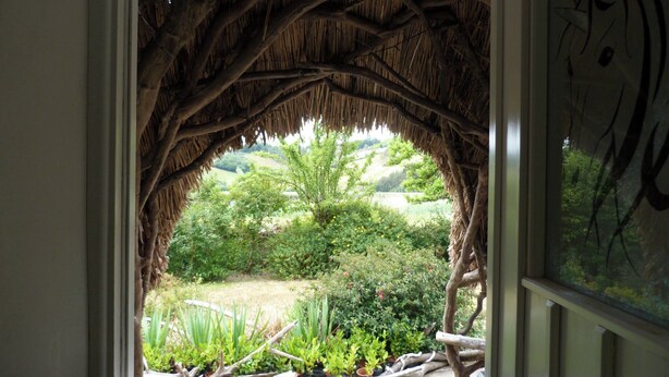 A window with a naturalistic frame made of branches and leaves, offering a view of a lush garden with various plants and trees.