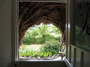 Une fenêtre ornée d’un cadre naturaliste fait de branches et de feuilles, qui offre une vue sur un jardin luxuriant, où poussent différentes plantes et arbres.