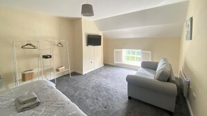 Une chambre à coucher avec un lit, un canapé gris, un téléviseur fixé au mur et une fenêtre avec des stores.