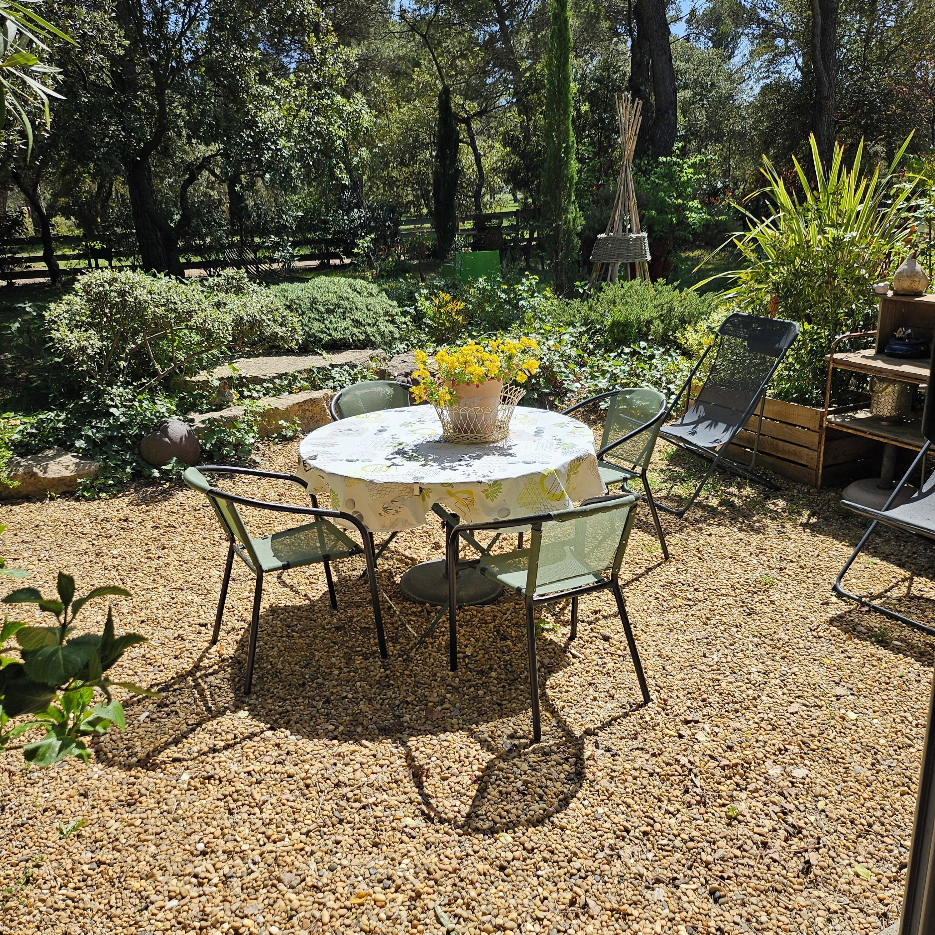 Une table ronde entourée de chaises, posée sur du gravier, dans un jardin où l’on trouve diverses plantes et où une clôture en bois se dresse à l’arrière-plan.