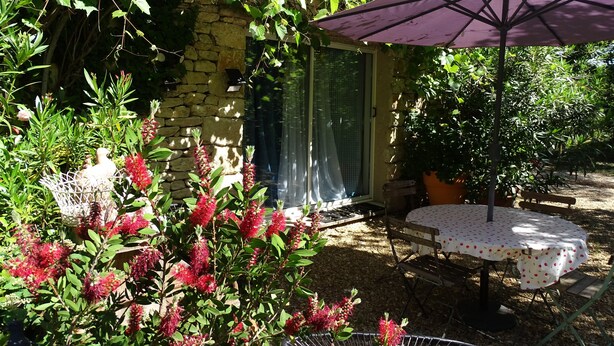 A garden with a stone wall, a table with a purple umbrella, and red flowering plants. - Chambre d hôte (Murs)
