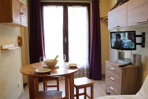 Quarto acolhedor com uma mesa de jantar de madeira posta para dois, uma televisão na parede e uma cama com edredom branco.