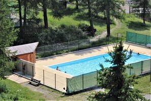 Ein rechteckiger Pool mit Holzboden und einem kleinen Holzgebäude, umgeben von Bäumen und einer Rasenfläche.