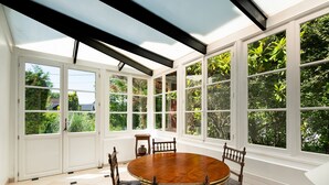Une véranda avec une grande table et des chaises en bois, des fenêtres à cadre blanc et une vue sur la verdure environnante.