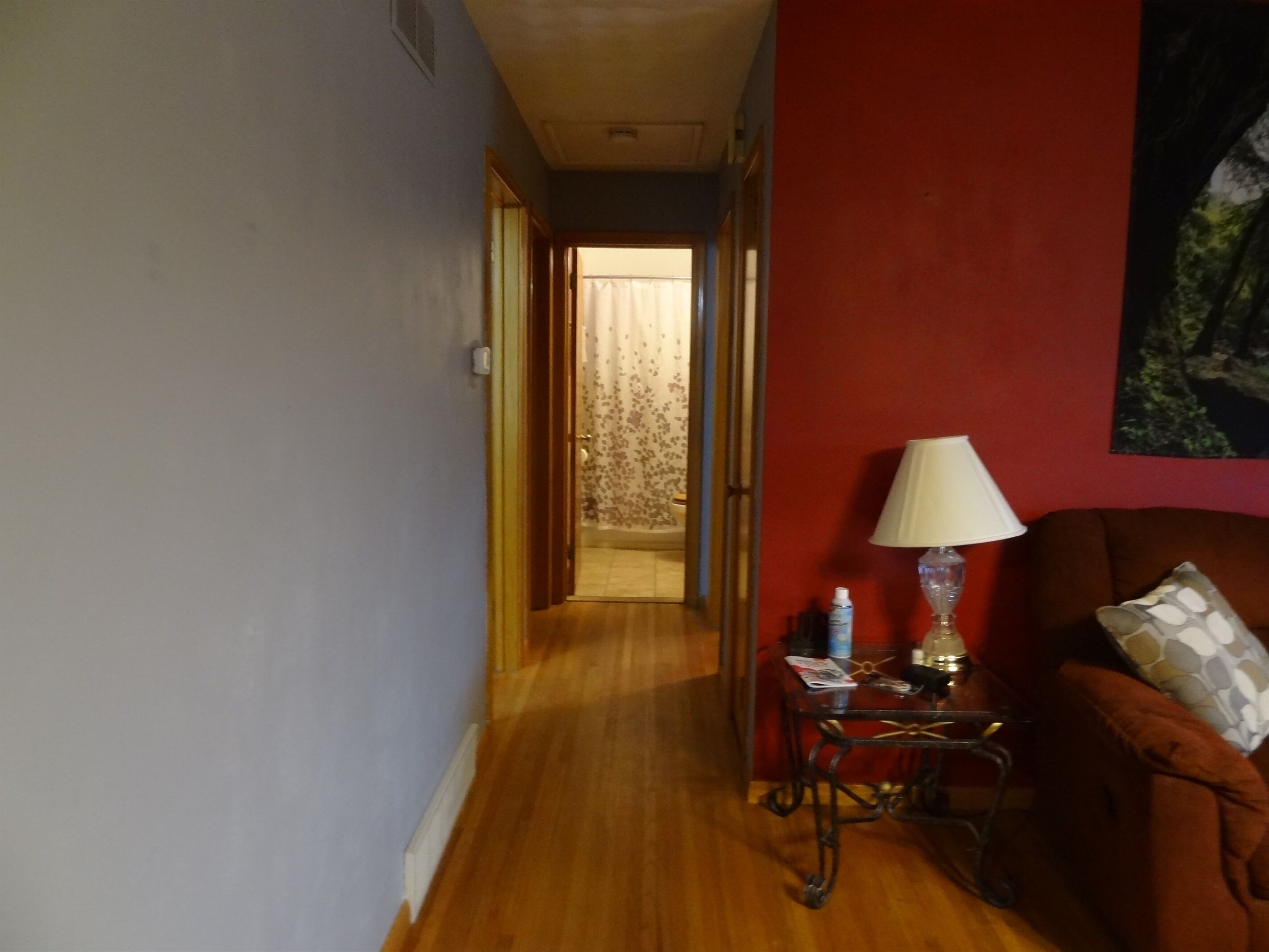 A hallway with a wooden floor, a red wall, and a door leading to another room. There is a table with a lamp and some items on it.