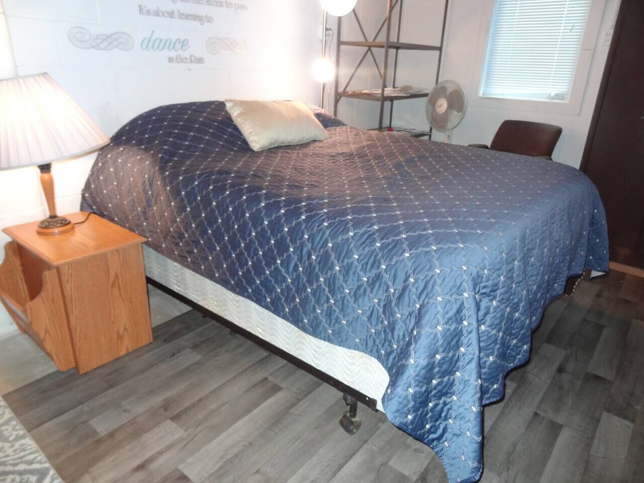 Une chambre à coucher avec un lit, une table de chevet, une lampe, un ventilateur et une fenêtre avec des stores.