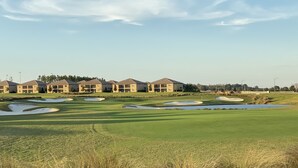 A golf course with bunkers, a water hazard, and a row of buildings in the background.