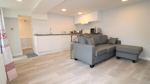 A modern living room with a grey sectional sofa, a kitchen area with white cabinets, and a wooden floor.