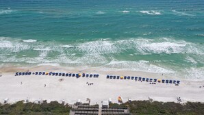 An aerial view of a beach with rows of umbrellas and lounge chairs, a sandy shore, and the ocean in the background.