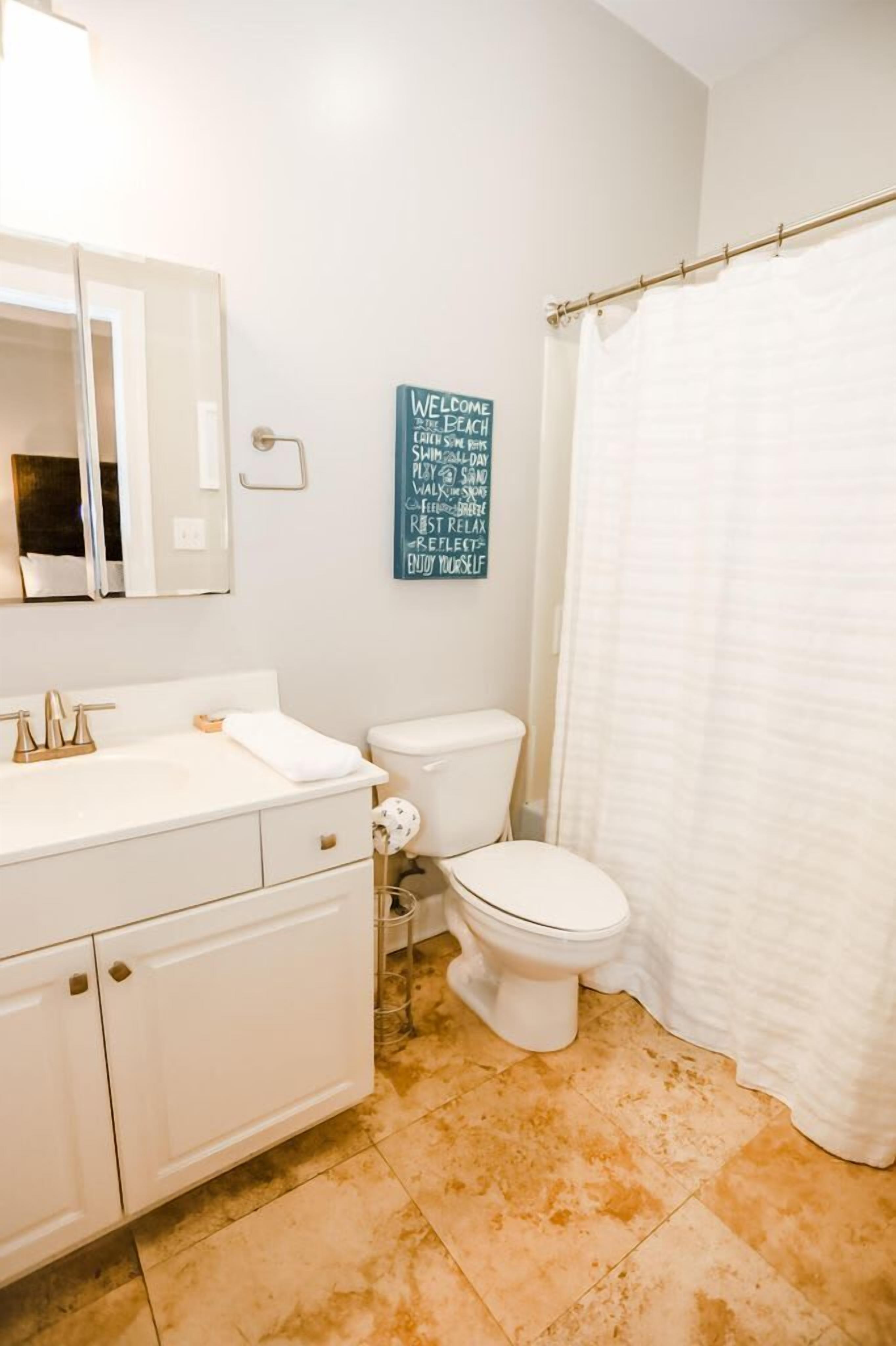 A bathroom with a white sink, mirror, and toilet. There is a shower curtain and a motivational sign on the wall.