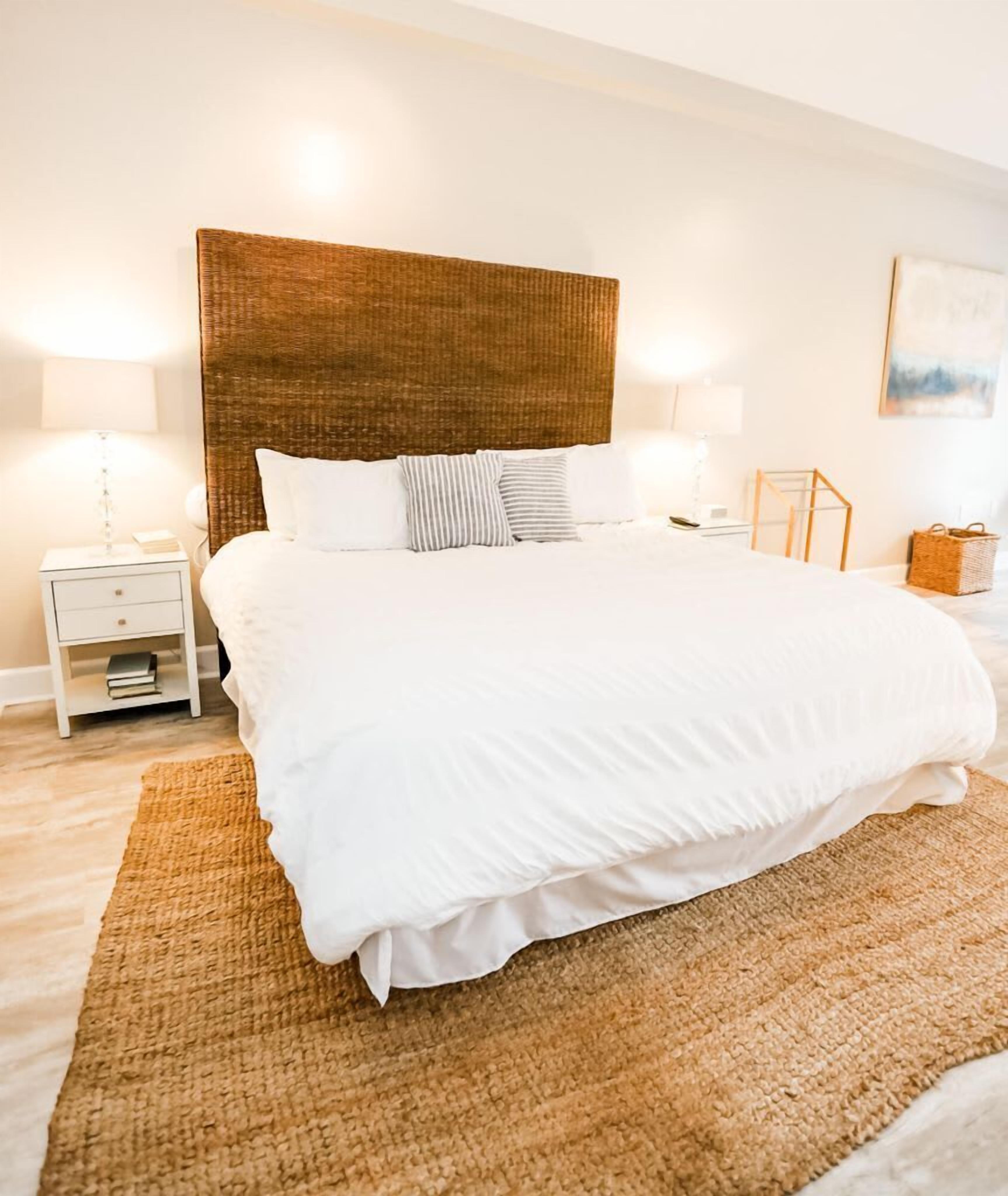 Una cama bien hecha con ropa blanca, un cabecero tejido y una alfombra con textura en el suelo.